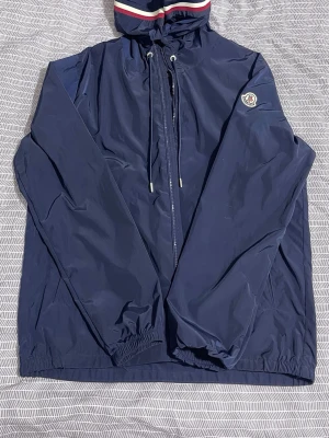 Marinblå vindjacka från Moncler - Snygg marinblå vindjacka från Moncler med huva och dragsko. Jackan har dragkedja framtill, elastiska muddar och Moncler-logga på ärmen. Huva med röd, vit och blå randig kant. Perfekt för blåsiga dagar och stilren look.