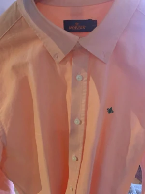 Persikofärgad skjorta från Morris - Säljer en persikofärgad skjorta från Morris med klassisk krage och knappar framtill. Skjortan har lång ärm och en liten grön broderad logga på bröstet. Tillverkad i bomull för en skön känsla och snygg passform.