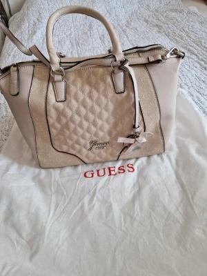 Beige handväska från Guess - Snygg beige handväska från Guess med quiltat mönster och ljusa detaljer. Väskan har dubbla handtag, avtagbar axelrem och dekorativ rosett. Metallogo framtill och dragkedja upptill. Perfekt för dig som gillar stilrena accessoarer.