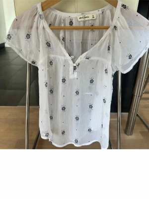 Vit blommig blus från Abercrombie - Söt vit blus från Abercrombie med små svarta blommor och kort ärm. Blusen har v-ringning med knytning och är gjord i ett tunt, luftigt tyg som känns perfekt för varma dagar. Lätt transparent och avslappnad passform.
