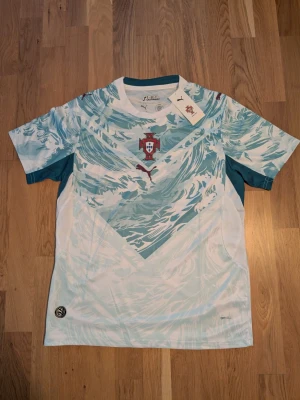 Portugal fotbollströja Puma blåvit. Storlek.S.M - Helt ny. Säljer en Portugal fotbollströja från Puma i blåvitt vågmönster. Tröjan har korta ärmar, rund hals och Portugal-emblem på bröstet. Tillverkad i lätt och ventilerande material, perfekt för träning eller match.