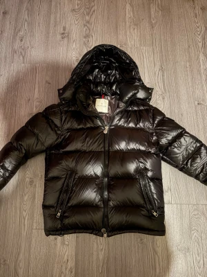 Svart dunjacka från Moncler med huva - Snygg svart dunjacka från Moncler med glansig finish och avtagbar huva. Jackan har två dragkedjeförsedda fickor framtill och quiltad design. Perfekt för kalla vinterdagar och riktigt varm tack vare dunfyllningen. OBS! Pris och byte kan diskuteras.
