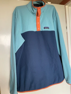 Patagonia fleece tröja blå/turkos - Säljer en snygg fleece tröja från Patagonia i turkos och marinblå med orange detaljer. Tröjan har hög krage, knappslå framtill och logga på bröstet. Perfekt för lager-på-lager och chill dagar.