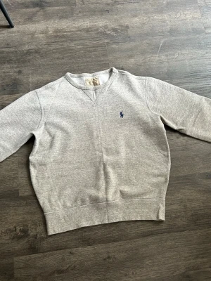 Grå sweatshirt från Ralph Lauren - Klassisk grå sweatshirt från Ralph Lauren med broderad blå logga på bröstet. Tröjan har rund halsringning, långa ärmar och ribbade muddar vid ärmslut och nederkant. Perfekt för en chill och stilren look.