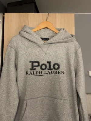 Grå hoodie från Polo Ralph Lauren - Snygg grå hoodie från Polo Ralph Lauren med stor svart logga framtill. Klassisk modell med huva och magficka, perfekt för en avslappnad stil. Tillverkad i mjukt material som känns skönt mot huden.