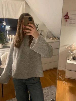 Grå oversized stickad tröja - Mysig grå stickad tröja med oversized passform och vida ärmar. Tröjan har en enkel design utan synliga detaljer eller mönster och är perfekt för lager-på-lager. Materialet ser ut att vara mjukt och fluffigt, troligen ullblandning.