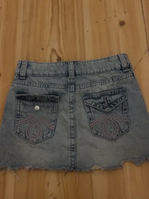 Grå jeanskjol med rå kant - Cool grå jeanskjol med rå, fransig kant och snygga bakfickor med broderi. Kjolen har bälteshällor, knapp och dragkedja framtill samt slitna detaljer för en trendig look. Perfekt för dig som gillar en avslappnad och edgy stil.