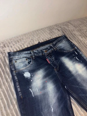 Mörkblå Dsquared2 ICON jeans - Snygga mörkblå jeans från Dsquared2 med slitningar, färgstänk och en röd ICON-detalj bak i midjan. Klassisk femficksmodell med raka ben och cool tvätt. Perfekta för dig som gillar streetstyle och vill sticka ut lite extra.