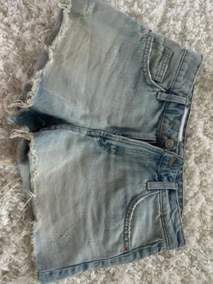 Ljusblå lågmidjade korta jeansshorts med fransig kant från Zara storlek 32 - Säljer ett par ljusblå jeansshorts med fransig kant och slitna detaljer. Låg midja med perfekt passform. Klassisk femficksmodell med dragkedja och knapp. Perfekta för sommaren och ger en avslappnad vibe till din outfit. Storlek 32 från Zara. Mycket fint skick!