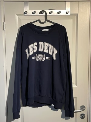 Mörkblå sweatshirt från Les Deux - Mörkblå sweatshirt från Les Deux med tryckt logga och text 'LES DEUX EST. 2011' på bröstet. Tröjan har rund halsringning, långa ärmar och ribbade muddar vid ärmslut och nederkant. Perfekt för en avslappnad och stilren look. Storlek xl men passar folk som har L 