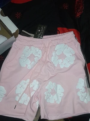 Denim tears - Säljer ett par ljusrosa shorts med vita blommönster och snörning i midjan. Shortsen har avslappnad passform och är perfekta för sommaren. Mjukt material och elastisk midja för extra komfort.