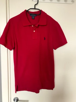 Röd pikétopp från Ralph Lauren - Röd pikétröja från Ralph Lauren med klassisk krage, två knappar och broderad blå logga på bröstet. Tillverkad i mjuk bomull och har korta ärmar. Enkel och stilren modell som funkar till det mesta. Skön och fin tröja som är lite extra stilig men ändå casual.