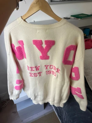 Vit stickad tröja med rosa NYC-tryck - Vit stickad tröja från Sisters Point med stora rosa bokstäver som bildar 'NYC' och texten 'New York Est 1992' framtill. Tröjan har rund halsringning, långa ärmar och ribbade muddar. Perfekt för dig som gillar streetstyle och statementplagg.
