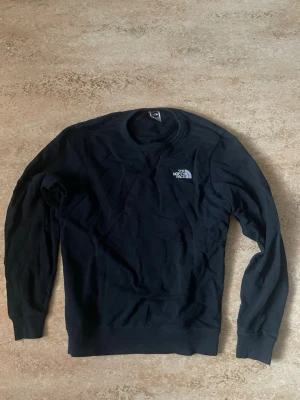 Svart sweatshirt från The North Face - Klassisk svart sweatshirt från The North Face med rund halsringning och logga på bröstet samt ryggen. Tröjan har långa ärmar och ribbade muddar vid ärmslut och nederkant. Storlek S.