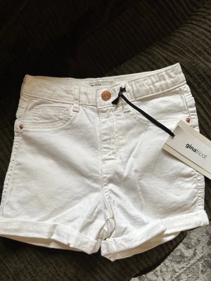 Vita high waist shorts Gina Tricot - Snygga vita shorts från Gina Tricot i klassisk jeansmodell med hög midja. De har uppvikta benslut, fem fickor och knappgylf. Perfekta till sommaren och lätta att matcha med olika toppar.