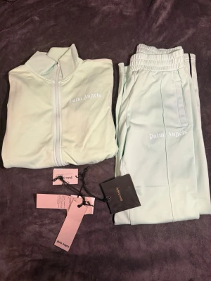 Mintgrönt loungewear-set Palm Angels - Superfräscht loungewear-set från Palm Angels i mintgrönt. Setet består av en ziptröja med hög krage och matchande byxor med resår i midjan. Båda delarna har Palm Angels-logga tryckt i vitt. Perfekt för chill dagar hemma eller när du vill vara bekväm men ändå snygg.
