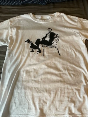 Vit t-shirt med svart tryck - Säljer en vit t-shirt med ett svart tryck av Ulf K. T-shirten har rund hals och korta ärmar. Perfekt för dig som gillar unika motiv och streetstyle.