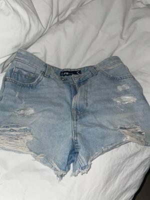Ljusa blå jeansshorts med slitningar - Säljer ett par ljusa blå jeansshorts från Faded med råa kanter och slitna detaljer framtill. Klassisk femficksmodell med hög midja och dragkedja. Perfekta för sommardagar och en avslappnad stil. Skriv för mer bilder