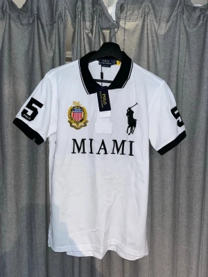 Vit pikétröja Polo Ralph Lauren Miami - Vit pikétröja från Polo Ralph Lauren med svart krage och svarta detaljer. Stor broderad logga, texten MIAMI och siffran 5 på ärmar och rygg. Klassisk polokrage och korta ärmar, perfekt för en sportig look.