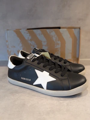 Svarta Golden Goose sneakers med stjärna - Svarta sneakers från Golden Goose med vit stjärna på sidan och vit häl. Skorna har svarta skosnören, GGDB-logga på plösen och en beige sula. Tillverkade i läder och har en klassisk låg profil som passar till streetwear.