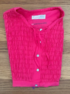 Rosa blus från Odd Molly storlek 2, M - Säljer en rosa blus från Odd Molly med smockad framsida, knappar och knytband vid halsen. Blusen har ett broderat fjärilsmärke på ryggen med texten 'Odd Molly Classic'. Tillverkad i mjukt bomullstyg. Är cerise, ej röd!