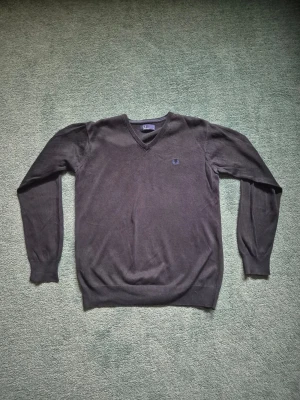 Blå V-ringad tröja Fred Perry - Klassisk svart V-ringad tröja från Fred Perry med diskret broderad logga på bröstet. Tröjan har långa ärmar och ribbade muddar vid ärmslut och nederkant. Perfekt för en stilren och enkel look.