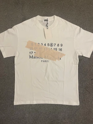 Vit Maison Margiela t-shirt med tryck - Vit t-shirt från Maison Margiela med ikoniskt siffertryck och beige tejpdetalj över bröstet. Klassisk passform och rund halsringning. Perfekt för dig som gillar streetstyle och unika designer.
