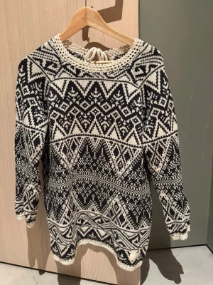 Svartvit stickad tröja från Odd Molly - Stickad Odd Molly tröja i svart och vitt med klassiskt nordiskt mönster. Tröjan har rund halsringning med virkad kant och knytning bak. Långärmad och mjuk, perfekt för kalla dagar. Passar dig som gillar retrovibbar och grafiska detaljer.