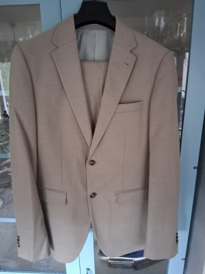 Beige kostym från Riley - Stilren beige kostym från Riley med tvåknäppt kavaj, klassiska slag och matchande byxor. Inköpt på Brothers för 2 år sedan, använd 1 gång på skolavslutning. Kavaj stlk 48, byxor stlk 50