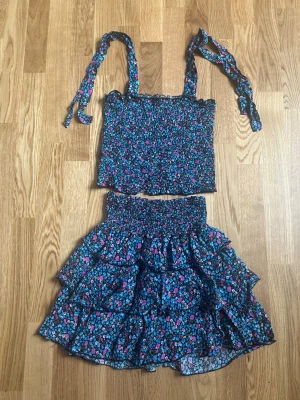 Blommigt tvådelat set med volanger - Sött tvådelat set med smockad topp och kjol i svart tyg med blått och rosa blommönster. Toppen har knytbara axelband och kjolen har bred smock i midjan samt flera lager volanger. Materialet känns lätt och luftigt, perfekt för varma dagar.