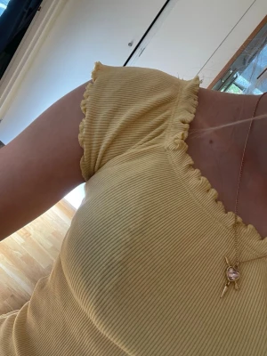 Gul ribbad topp med volangkant - Söt gul ribbad topp med korta ärmar och volangkant vid halsringning, ärmslut och nederkant. Toppen är croppad och har en figurnära passform, perfekt för en somrig look.