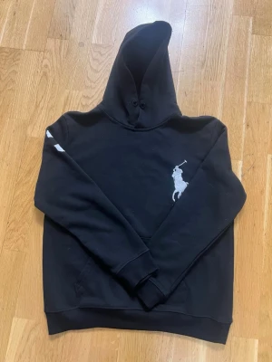 Svart hoodie från Polo Ralph Lauren - Snygg svart hoodie från Polo Ralph Lauren med stor vit broderad logga på bröstet. Klassisk passform med huva och ribbade muddar. Tillverkad i mjukt bomullsmaterial som känns skönt mot huden. Perfekt för en avslappnad och stilren look.