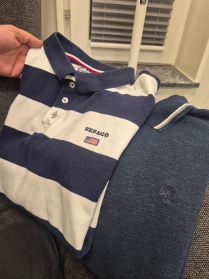 Två pikétröjor från Sebago o Vintage skill L Xl - Säljer två pikétröjor från Sebago. Ena är blåvit med breda ränder, broderad logga och amerikansk flagga på bröstet. Den andra är marinblå med vit detalj på kragen och diskret logga. Båda har klassisk pikékrage och knappar framtill. Både är samma storlek passar större L eller mindre XL