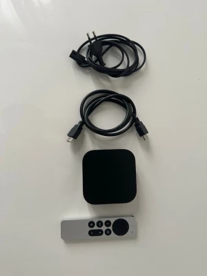 Apple TV 4K med fjärrkontroll och kablar - Apple TV 4K mediaspelare i svart med tillhörande silverfärgad fjärrkontroll, strömkabel och HDMI-kabel. Perfekt för streaming av filmer, serier och appar direkt till din TV.