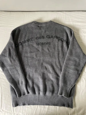 Grå stickad tröja Comme des Garçons - Säljer en grå ribbstickad tröja från Comme des Garçons Homme. Tröjan har rund halsringning och tryckt logga på ryggen. Perfekt för dig som gillar minimalistisk och stilren design.