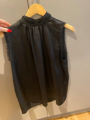 Svart ärmlös blus med hög krage - Säljer en svart ärmlös blus med hög krage och råa kanter. Blusen har en lätt transparent känsla och ett mjukt, flytande tyg som ger en elegant look. Perfekt för dig som gillar stilrena och trendiga plagg.