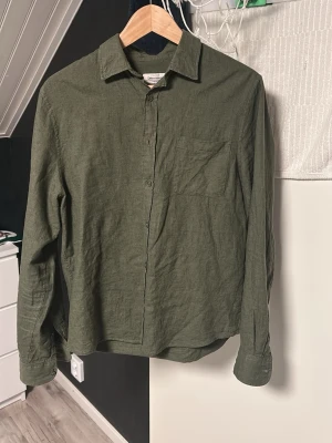 Olivgrön linneskjorta från Dressman - Stilren olivgrön skjorta i linne från Dressman. Skjortan har klassisk krage, bröstficka och knappar framtill. Perfekt för en avslappnad men snygg look. Långärmad och med en loose passform.