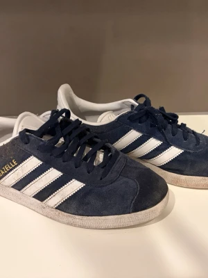 Adidas Gazelle blå och vita sneakers - Klassiska Adidas Gazelle sneakers i mörkblå mocka med vita detaljer och de ikoniska tre ränderna på sidorna. Skorna har vit sula och vit plös med Adidas-logga. Perfekta för en stilren och sportig look.