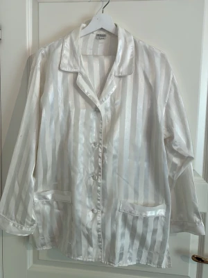 Pyjamaset i satinliknande tyg - Snyggt vitt pyjamasset med blanka vertikala ränder. Klassisk krage, knäppning framtill och två fickor. Perfekt för en chill kväll hemma eller när du vill känna dig extra lyxig. Långärmad och mjuk mot huden.