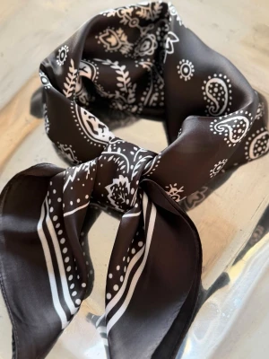 Midnight Paisley Scarf 70x70 – Satin Feeling - Elegant scarf i djup svart med vita paisleydetaljer och satinlik finish. En tidlös accessoar med mjukt fall och klassisk bandana-inspirerad känsla som passar både dam och herr. Fin runt halsen, i håret eller knuten på väskan.  • 70 x 70 cm • Satin feeling med mjukt lyster • Svart/vit paisleydesign • Unisex • Limited Edition