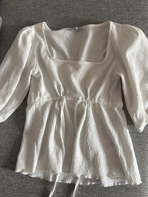 Vit blus med fyrkantig ringning - Stilren vit blus med fyrkantig ringning och trekvartsärm. Blusen har en markerad midja med knytband och ett lätt strukturerat tyg som ger en luftig känsla. Perfekt att matcha med jeans eller kjol för en trendig look.