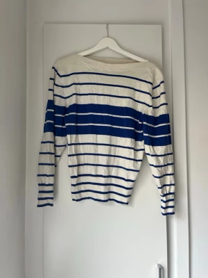 Randig tröja från GANT i blått och vitt - Snygg långärmad tröja från GANT i vitt med blåa horisontella ränder. Tröjan har en klassisk båtringad hals och är tillverkad i ett mjukt material som känns skönt mot huden. Perfekt för en stilren och avslappnad look.