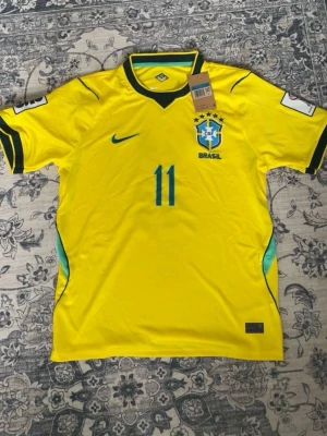 Brasilien matchtröja Raphinha #11 - Gul Brasilien fotbollströja från Nike med nummer 11 och namnet Raphinha på ryggen. Tröjan har blå detaljer, CBF-emblem på bröstet och svarta ränder på ärmarna. Perfekt för dig som älskar landslagsfotboll och vill visa ditt stöd.
