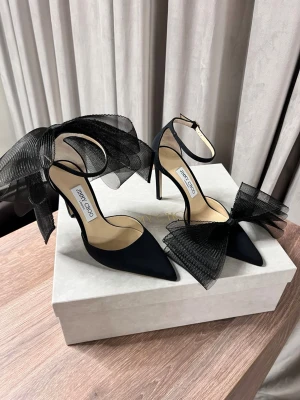 Svarta pumps med rosett från Jimmy Choo - Stilrena svarta pumps från Jimmy Choo med spetsig tå, hög klack och stor dekorativ rosett i mesh. Skorna har en elegant ankelrem med spänne och är tillverkade i ett matt tygmaterial. Perfekta för dig som vill sticka ut med en lyxig och trendig look.
