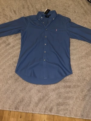 Blå skjorta från Ralph Lauren - Snygg blå skjorta från Ralph Lauren med klassisk button-down krage och den ikoniska rosa logotypen broderad på bröstet. Skjortan har långa ärmar, knappar framtill och är tillverkad i mjuk bomull. Perfekt för en stilren look.