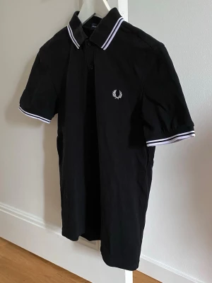 Fred Perry pikétröja  - Svart pikétröja från Fred Perry med vita ränder på krage och ärmslut. Storlek S. Nytvättad och i bra använt skick!