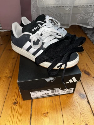 Adidas Grand Court Base 00s sneakers - Minimalt använda, snygga Adidas Grand Court Base 00s sneakers i svart mocka med vita detaljer, oanvända svarta snören följer med! Inköpta för 4 veckor sedan för priset 954kr, skorna 899kr + butiks impregnering 55kr. Säljes då jag tragiskt nog åkt på hälsporre och ej bör använda platta skor🙃