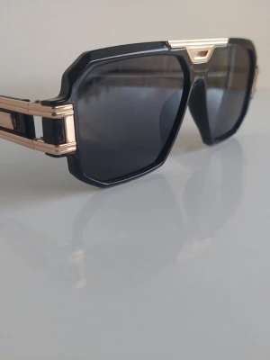 Svarta solglasögon med guldiga detaljer - Snygga solglasögon med svart båge och markerade guldiga detaljer på sidorna. Glasen är mörka och bågen har en kantig, modern form som ger en cool look. Perfekta för att lyfta din stil under soliga dagar.