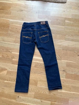 Mörkblå jeans från Nudie Jeans - Snygga mörkblå jeans från Nudie Jeans med klassisk rak passform och kontrastsömmar. Bakfickorna har Nudie Jeans ikoniska broderi. Perfekta för en stilren och avslappnad look. 30/32