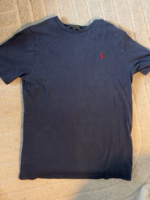 Polo Ralph Lauren T-shirt - Mörkblå t-shirt från Polo Ralph Lauren med klassisk passform och rund hals. T-shirten har en liten röd broderad logga på bröstet och är tillverkad i mjuk bomull. Perfekt för en stilren och avslappnad look. Tröjan är rätt så använd så säljs därav super billigt den är i storlek XL 18-20 så skulle säga att den sitter som en herr M